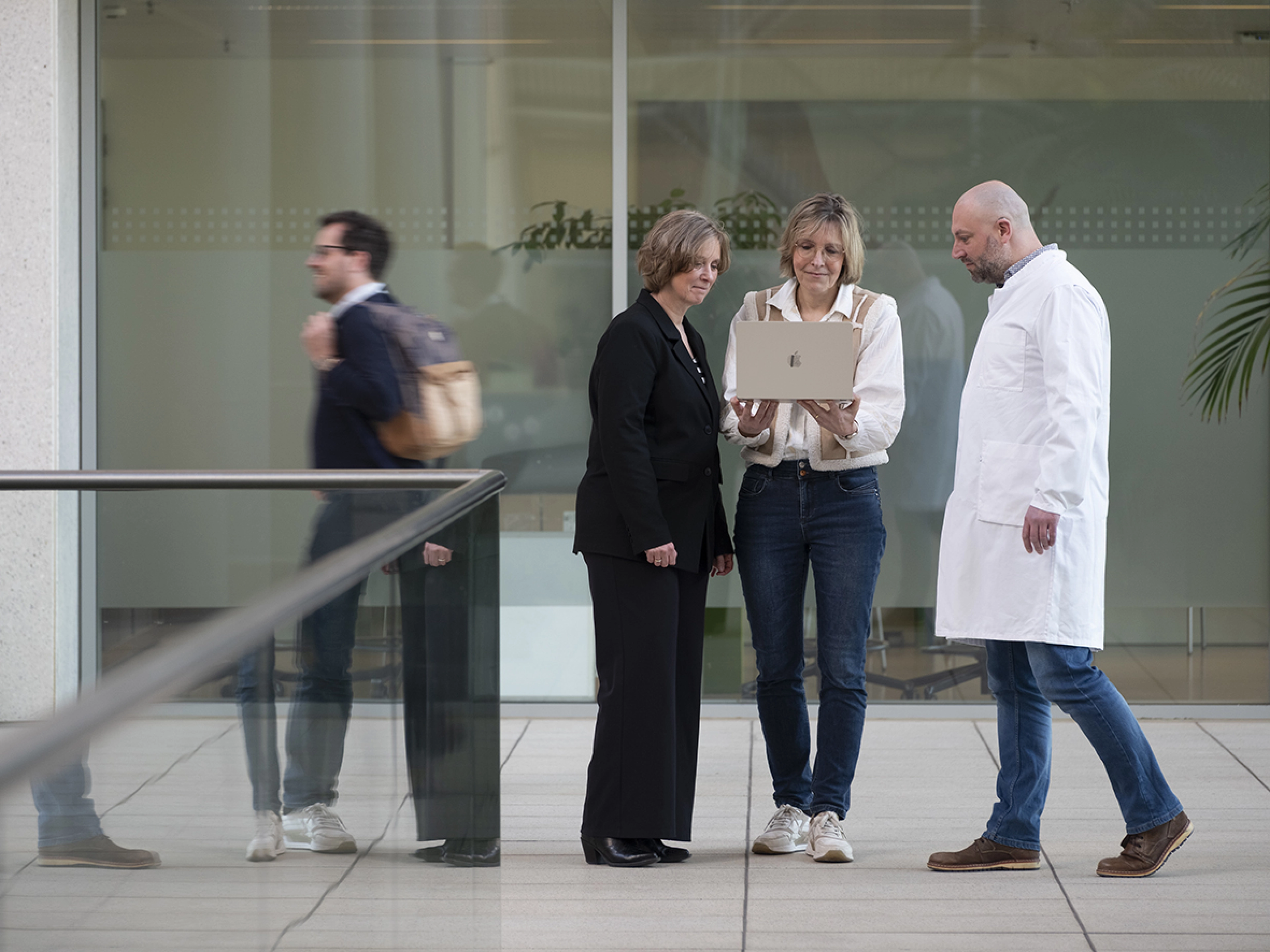 One man in a white coat and two women discussing and looking at a laptop. A man with backpack passing in the background.