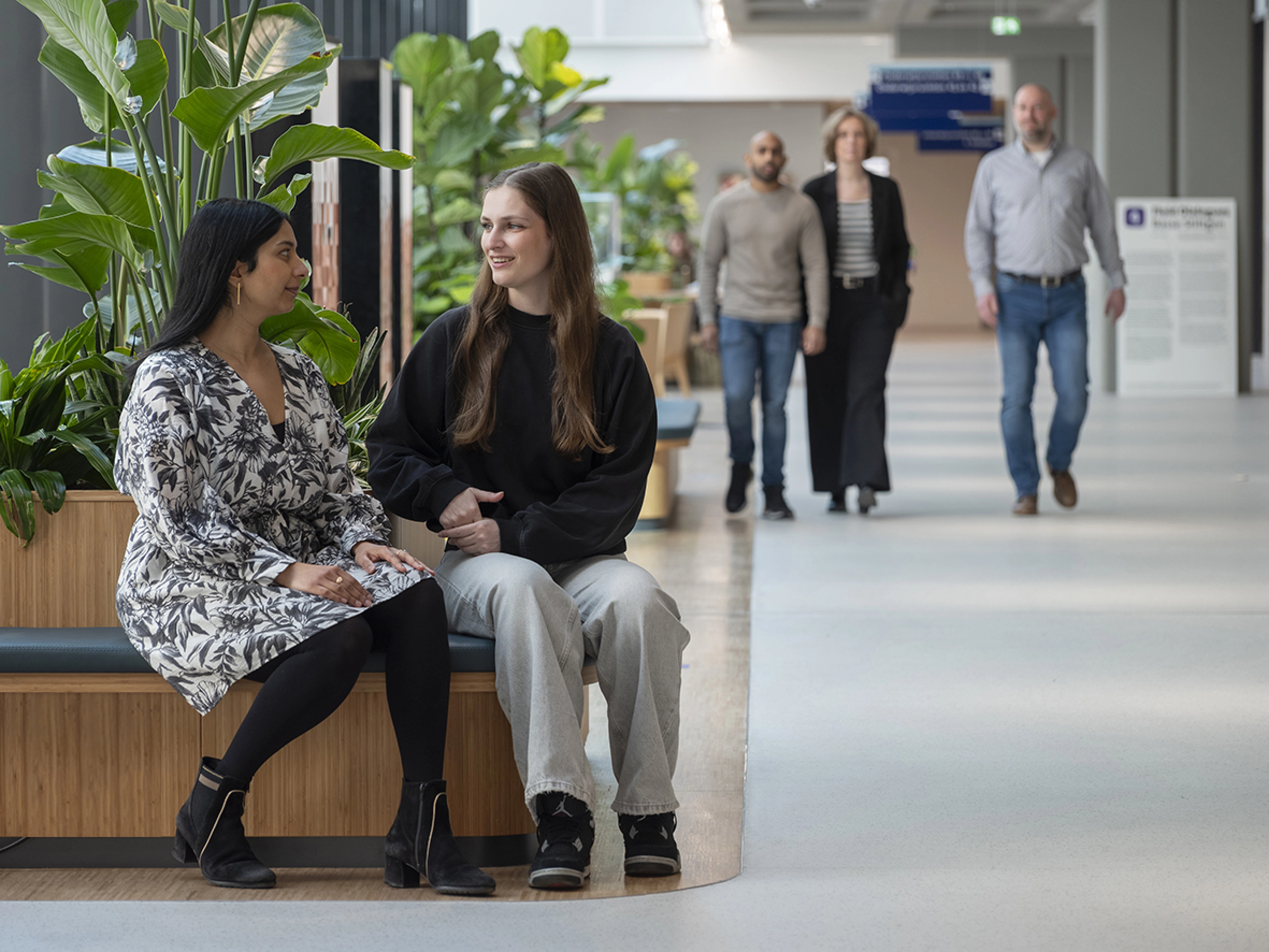 Two women sitting on a bench at the forfront, two man an a woman walking towards the viewer in the background.