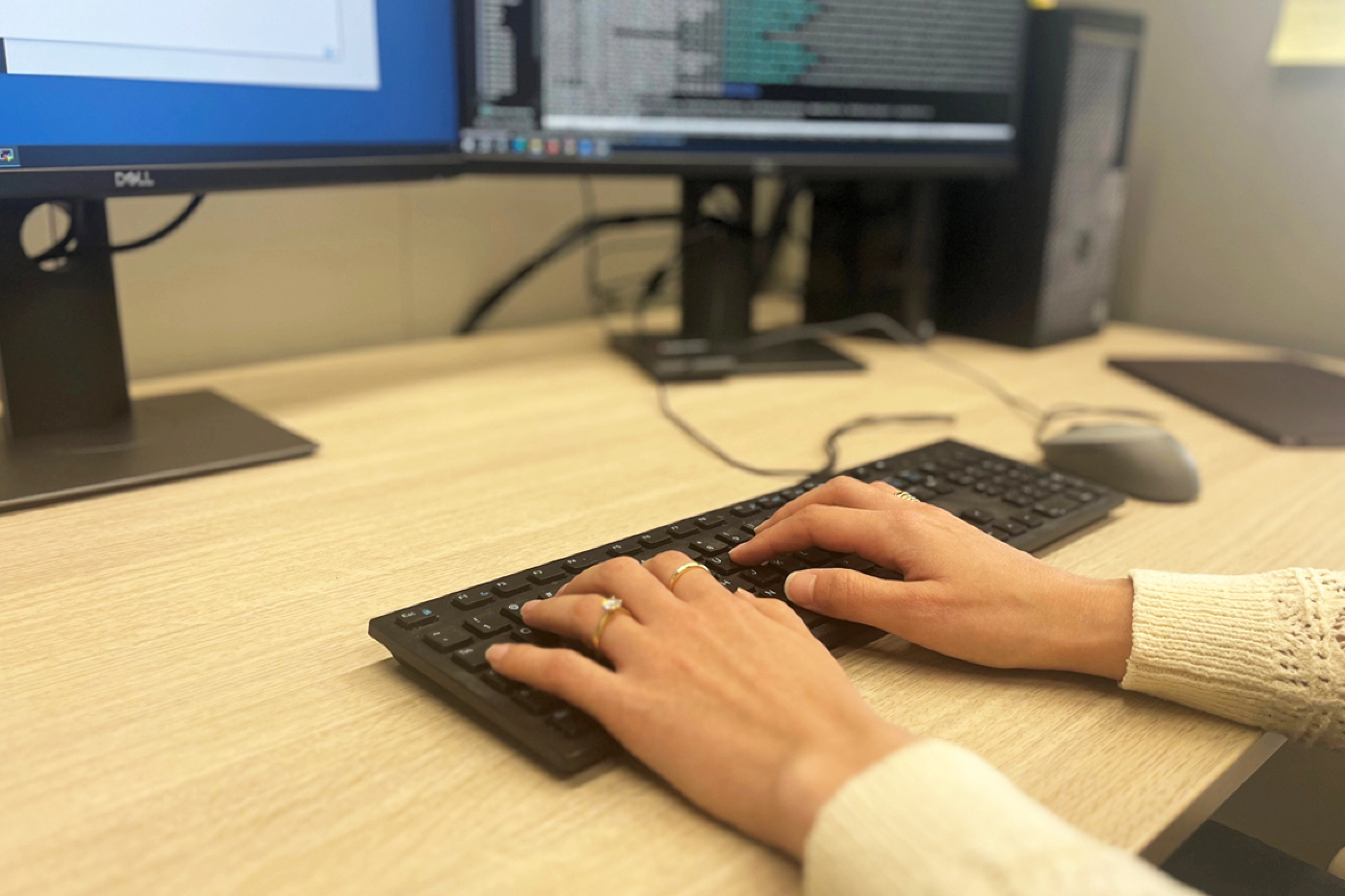 Two female hands on a black keyboard of a computer. Two computer screens in the background.