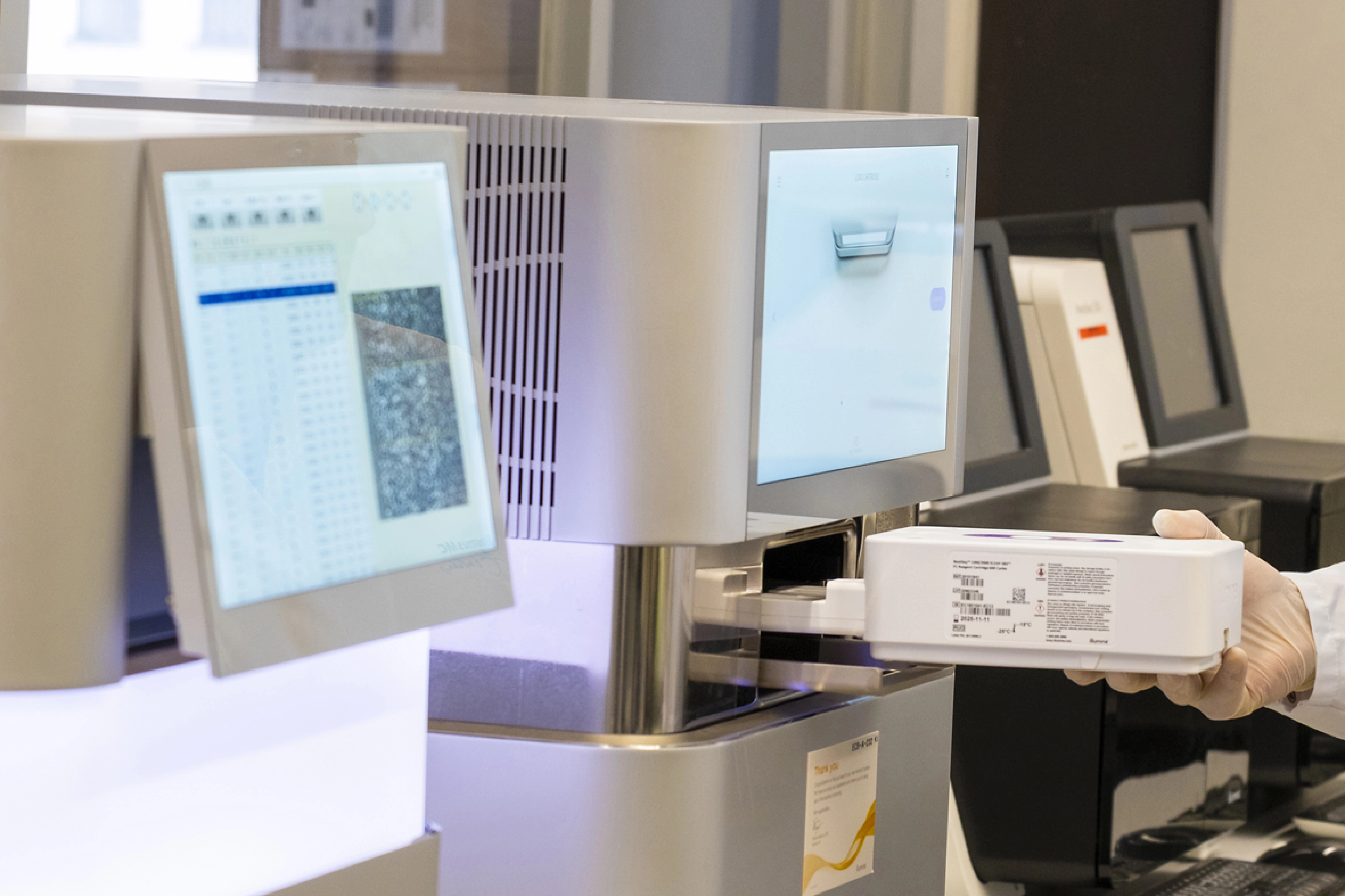 Sequencers seen from the side, with gloved hand of a technician working in a laboratory.